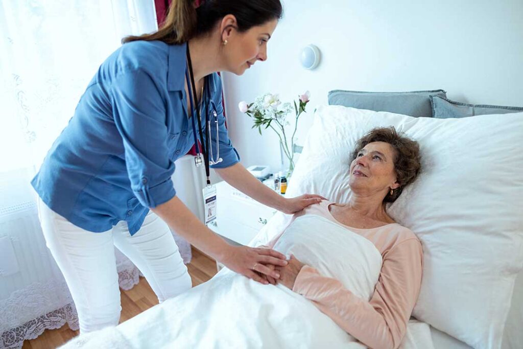 An elderly woman receives medication assistance from a caregiver in a warmly lit communal living room.