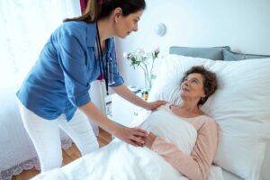 An elderly woman receives medication assistance from a caregiver in a warmly lit communal living room.